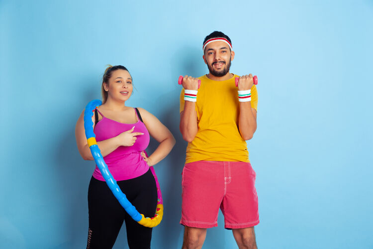手臂年轻漂亮的白人夫妇穿着鲜艳的衣服 在蓝色背景下训练运动概念 人类情感 表达 健康的生活方式 关系 家庭练习篮球和举重举行人护理