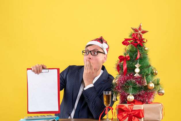 执行前视图惊奇的人拿着剪贴板坐在圣诞树旁边的桌子上 呈现在黄色背景上人树惊奇的人