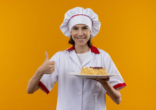 厨师身着厨师制服 面带微笑的白人年轻厨师女孩把蛋糕放在盘子里 竖起大拇指 在橙色背景上 留有复制空间蛋糕年轻盘子