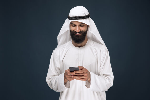 文化深蓝色工作室背景上的阿拉伯沙特男子半身肖像年轻男模使用智能手机 聊天商业 金融 面部表情 人类情感 技术的概念沙特反应严肃