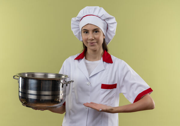 烹饪身着厨师制服的年轻高加索厨师女孩高兴地拿着并指着绿色背景上有复制空间的孤立锅锅制服女孩