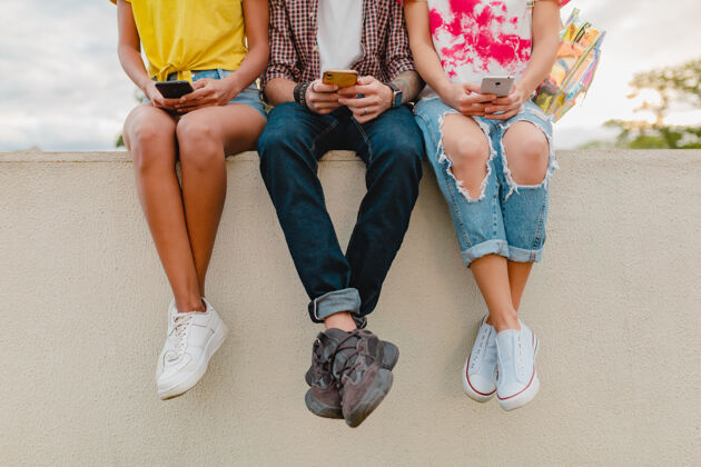 通信腿在运动鞋的年轻朋友公司坐在公园使用智能手机学生群组耳机