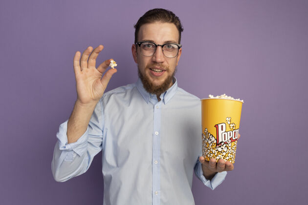脸一个戴眼镜的金发帅哥把一桶爆米花孤立地放在紫色的墙上人水桶男人