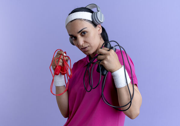 耳机年轻自信的白人运动女性戴着头带 戴着耳机戴着腕带 手里拿着跳绳 紫色背景上有复制空间头带佩戴腕带