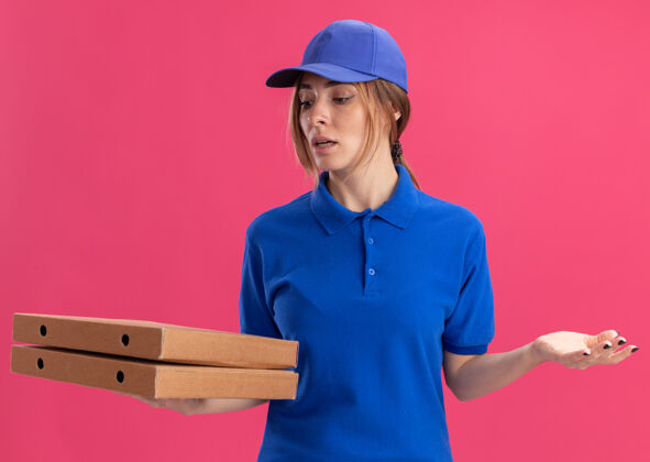 盒子困惑的年轻漂亮的女送货员在制服举行 并看着比萨饼盒隔离在粉红色的墙壁漂亮公民衣服
