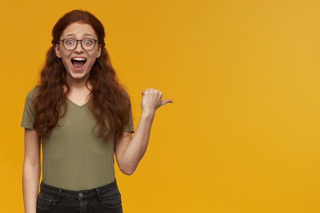 女人兴奋 惊讶的女人长着姜黄色的头发穿着绿色的t恤和眼镜用拇指指向右边的复印空间 隔离在橙色的墙上肖像年轻人服装