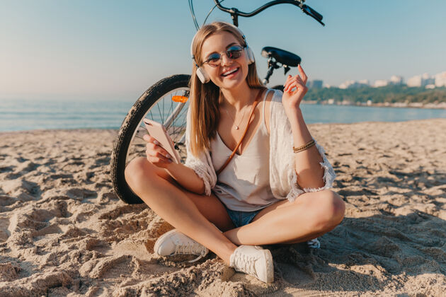 活跃时尚年轻迷人的金发微笑的女人坐在沙滩上骑着自行车戴着耳机听音乐休闲年轻配饰