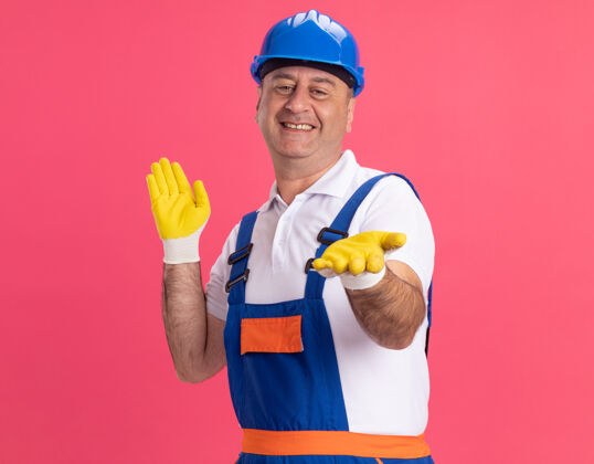 人微笑的成年建筑工人穿着制服 戴着防护手套 双手张开 孤立地站在粉红色的墙上公民制服保护
