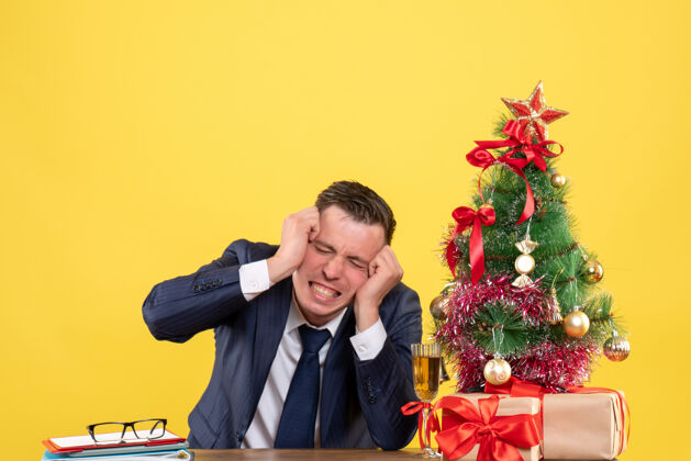 男正面图愤怒的男人抱着头坐在圣诞树旁的桌子旁 黄色背景上摆着礼物生意人抱着圣诞节