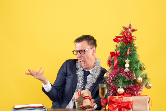 生意前视图愤怒的人戴着眼镜坐在圣诞树旁的桌子旁 黄色背景的礼物树桌子坐着