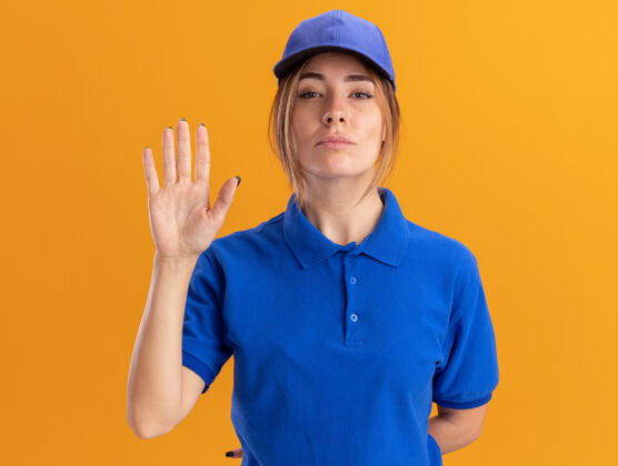 姿势自信的年轻漂亮的女送货员穿着制服站在橙色的墙上 举手孤立手脸人