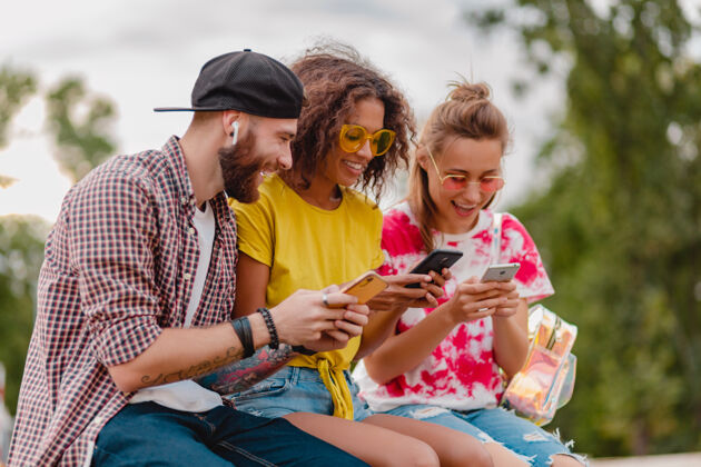 女人快乐的年轻朋友坐在公园里用智能手机微笑公司年轻时髦学生