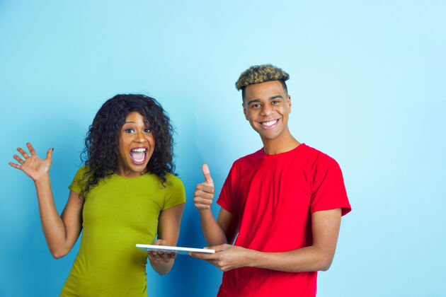 人类手持平板电脑 竖起大拇指年轻情绪化的非洲裔男女 穿着蓝色背景的彩色休闲服美丽的情侣人类情感的概念 面部表情 关系 广告一起男性头发