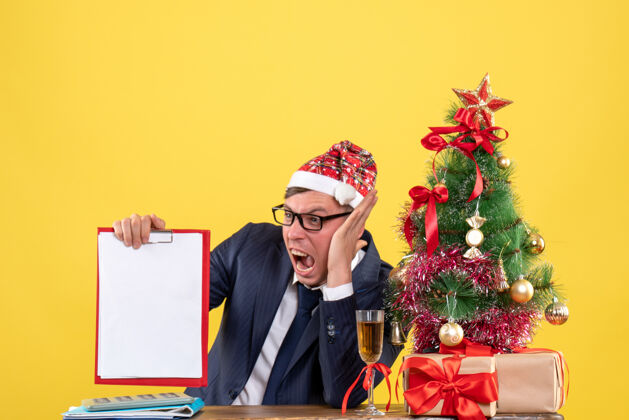 前面前视图愤怒的商人坐在圣诞树旁的桌子旁 呈现黄色背景近礼物圣诞节