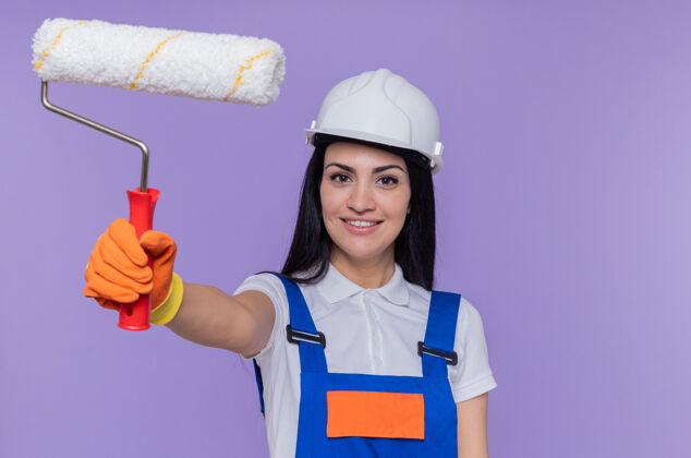 手握年轻的女建筑工人穿着施工制服 戴着安全帽 戴着橡胶手套 拿着油漆辊油漆站立年轻人