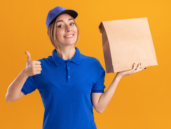 年轻身着制服 面带微笑的年轻漂亮女送货员竖起大拇指 把纸包裹隔离在橙色的墙上脸制服漂亮