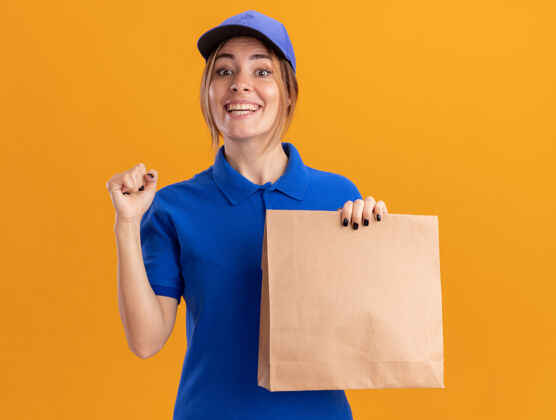 保持身着制服的年轻漂亮的女送货员微笑着握紧拳头 把纸包裹隔离在橙色的墙上抱着站着女孩