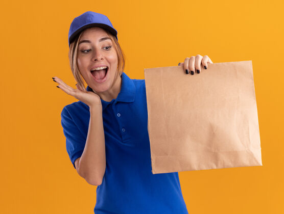 姿势穿着制服的年轻漂亮的女送货员兴奋地拿着纸包 看着隔离在橙色墙上的纸包包装漂亮女孩