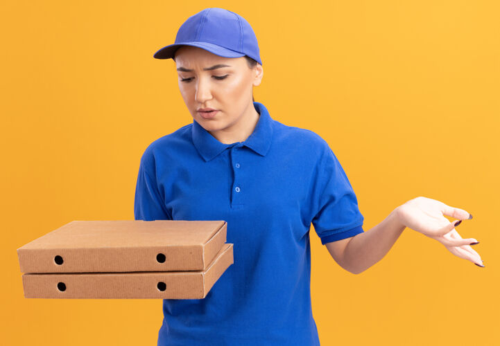 女人身穿蓝色制服 头戴鸭舌帽 拿着比萨饼盒的年轻女送货员站在橙色的墙上 看着他们困惑不解困惑年轻拿着