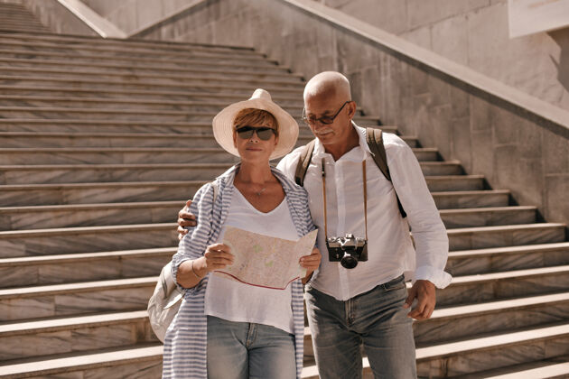 旅行戴着眼镜 穿着白衬衫和牛仔裤 拿着相机的时髦男人抱着戴着帽子的穿条纹衬衫的妻子可爱女士帽子