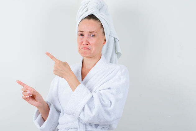 深色年轻的女士指着左上角的白色浴衣 毛巾和失望的神情 前视图完美女性女人
