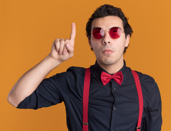 手指戴着眼镜 系着吊带 打着领结的时髦男人抬起头 困惑地用食指指着站在橙色墙上的东西吊带穿领带