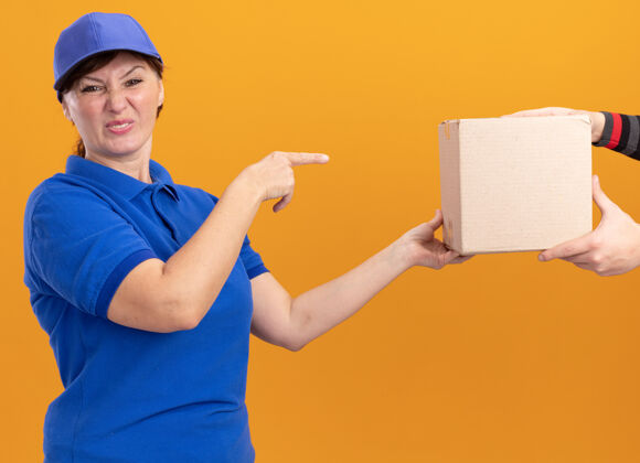 接收身着蓝色制服 戴着帽子的年轻送货员在收包裹时感到困惑 食指指着站在橙色墙上的盒子立场目录女人