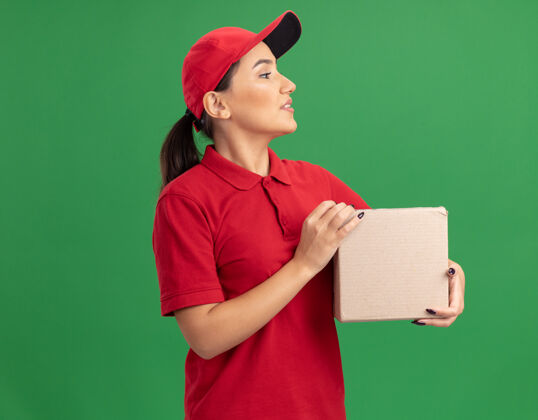 女人年轻的送货员身穿红色制服 头戴鸭舌帽 手拿纸板箱 面带微笑地站在绿色的墙边制服脸帽子