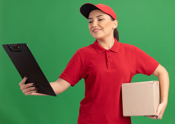 举行年轻的送货员身穿红色制服 头戴鸭舌帽 手里拿着一个纸板箱 手里拿着剪贴板 面带微笑地看着它站在绿色的墙上盒子立场微笑