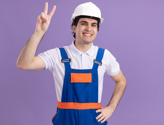手势身穿施工制服 头戴安全帽的年轻建筑工人面带微笑地看着前方 紫色的墙上挂着v字标志站立微笑建设者
