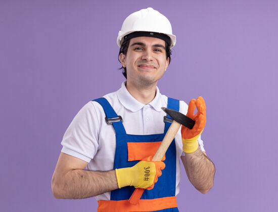 手套年轻的建筑工人穿着建筑制服 戴着安全帽 戴着橡胶手套 拿着锤子 面带微笑地站在紫色的墙上看着前方脸建筑站着
