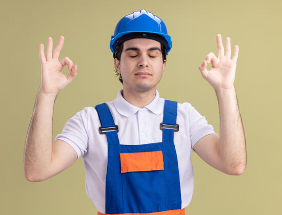 手指年轻的建筑工人穿着建筑制服 戴着安全帽 闭着眼睛站在绿色的墙上做着冥想的手势头盔制服制造