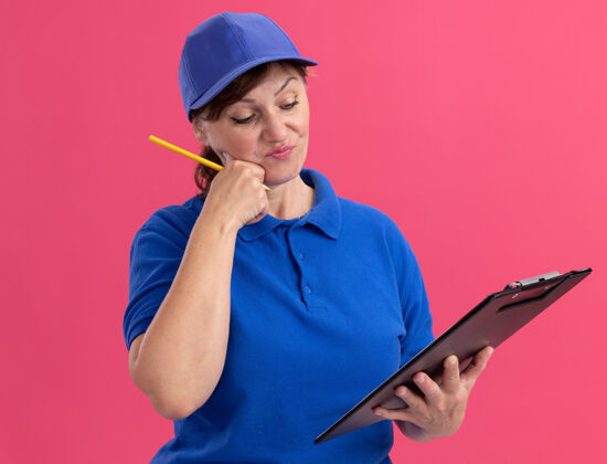 拿着身穿蓝色制服 头戴鸭舌帽的中年女送货员拿着剪贴板和铅笔站在粉色的墙上困惑地看着它帽子快递制服