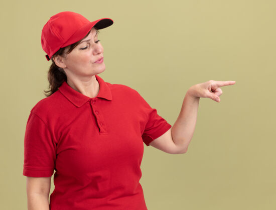 分娩身穿红色制服 头戴鸭舌帽的中年女送货员面带严肃的表情 食指指着站在绿色墙上的一边目录帽子脸