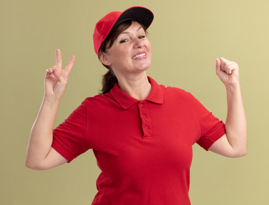 显示身穿红色制服 头戴鸭舌帽的中年女送货员站在绿色的墙上 面带微笑 脸上露出v字 举起拳头Vsign微笑帽子