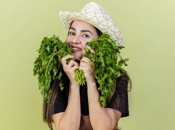 制服微笑美丽的园丁女孩穿着制服戴着园艺帽把香菜放在脸颊上孤立的橄榄绿背景女孩香菜微笑