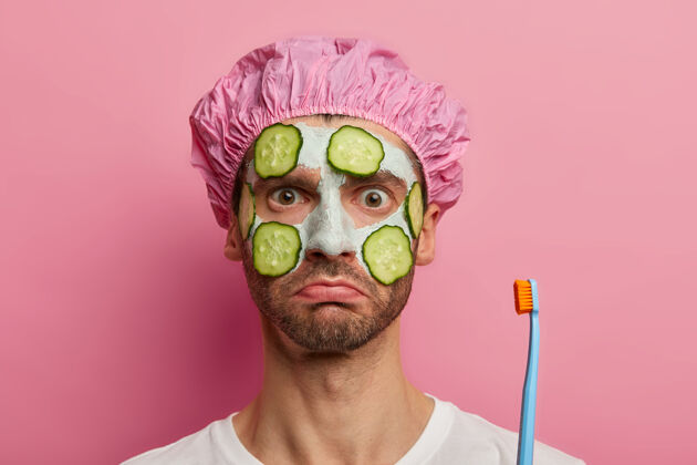 惊喜震惊男人的头像清洁面部皮肤 拿着牙刷 淋浴帽 准备清洁牙齿 有严肃严谨的造型 模特对抗粉色空间光滑奇迹T恤
