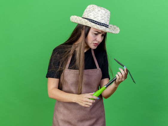 困惑迷茫美丽的园丁女孩穿着制服戴着园艺帽拿着锄头看耙子孤立在绿色的背景上持有制服穿