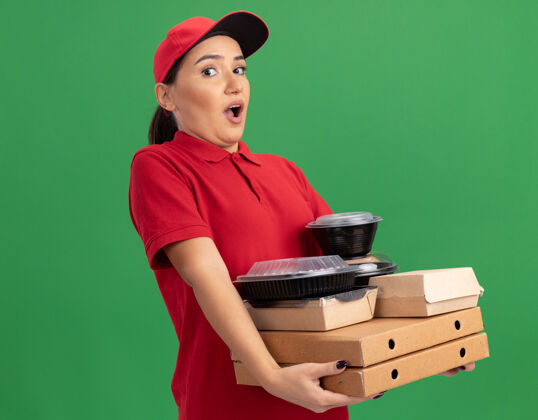 惊喜年轻的送货员身穿红色制服 头戴鸭舌帽 手里拿着披萨盒和食品包 站在绿色的墙边惊讶地看着前面女人站着送货