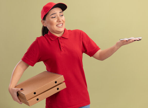 拿着身穿红色制服 头戴鸭舌帽 手拿比萨饼盒的年轻送货员站在绿色墙壁上 神情困惑地看着手中的智能手机移动帽子站着