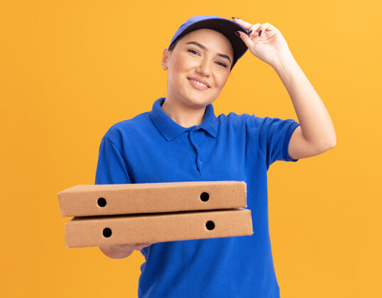 女人快乐的年轻送货员身穿蓝色制服 戴着帽子 手里拿着比萨饼盒 微笑着自信地站在橙色的墙上看着前面披萨盒子抱着