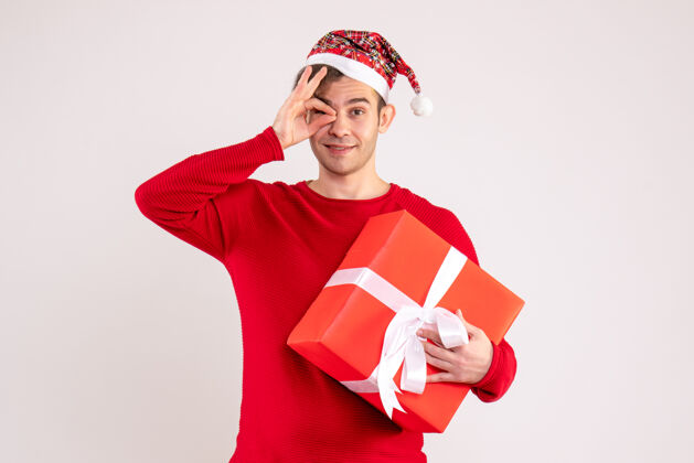 前面前视图：戴着圣诞帽的年轻人在白色背景上的一只眼睛前面放上了okey标志圣诞老人快乐礼物