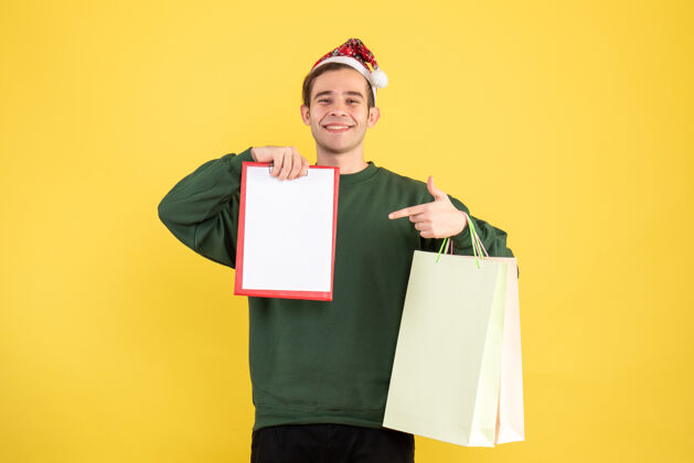 年轻人正面图：戴着圣诞帽的年轻人拿着购物袋和剪贴板站在黄色的背景复制空间上人帽子剪贴板