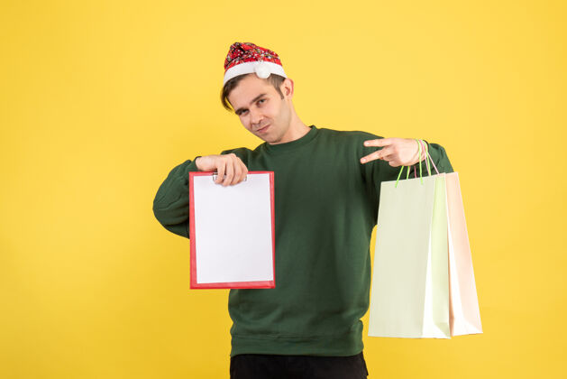 男人正面图：戴着圣诞帽的年轻人拿着购物袋和剪贴板站在黄色背景上圣诞节抱圣诞老人
