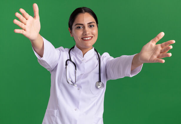 听诊器年轻的女医生 穿着医用外套 脖子上戴着听诊器 面带微笑 双手张开 站在绿色的墙上 做着欢迎的手势医学使外套