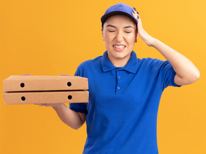 盒子身穿蓝色制服 头戴鸭舌帽 拿着比萨饼盒的年轻女送货员看起来很困惑 因为她站在橘色的墙上时犯了个错误帽子制服披萨