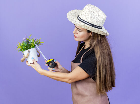 测量想着美丽的园丁女孩穿着制服戴着园艺帽测量花盆里的花用卷尺隔离在蓝色背景上思想花园艺