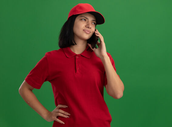 困惑站在绿色的墙上 穿着红色制服 戴着帽子的年轻送货员一边用手机说话 一边显得困惑和焦虑送货电话制服