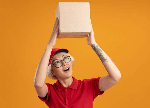 穿快乐的年轻送货员 身穿红色制服 戴着帽子 戴着眼镜 头上举着纸板箱 站在橙色的墙上 面带微笑地看着它制服微笑交货