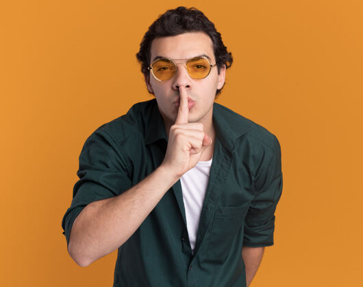 手势一个穿着绿衬衫戴着眼镜的年轻人站在橘色的墙上 手指放在嘴唇上做着沉默的手势年轻人嘴唇沉默